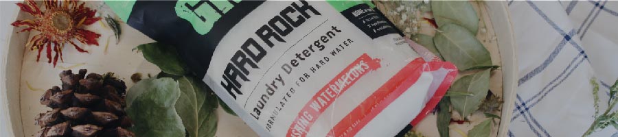 A bag of HARD ROCK laundry detergent surrounded by pinecones, dried flowers, and green leaves.