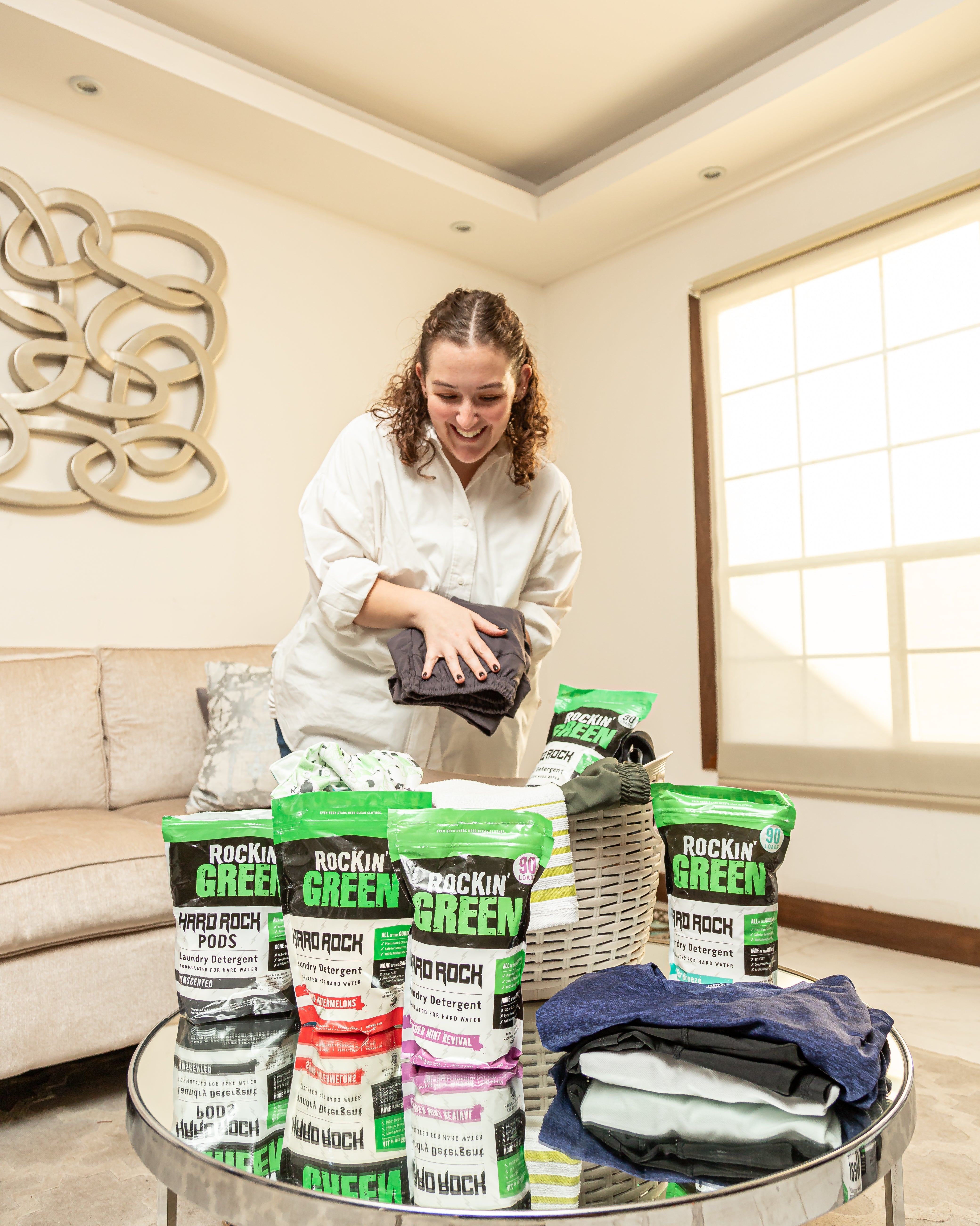 Woman folding laundry with Rockin Green products on a table in a bright living room.