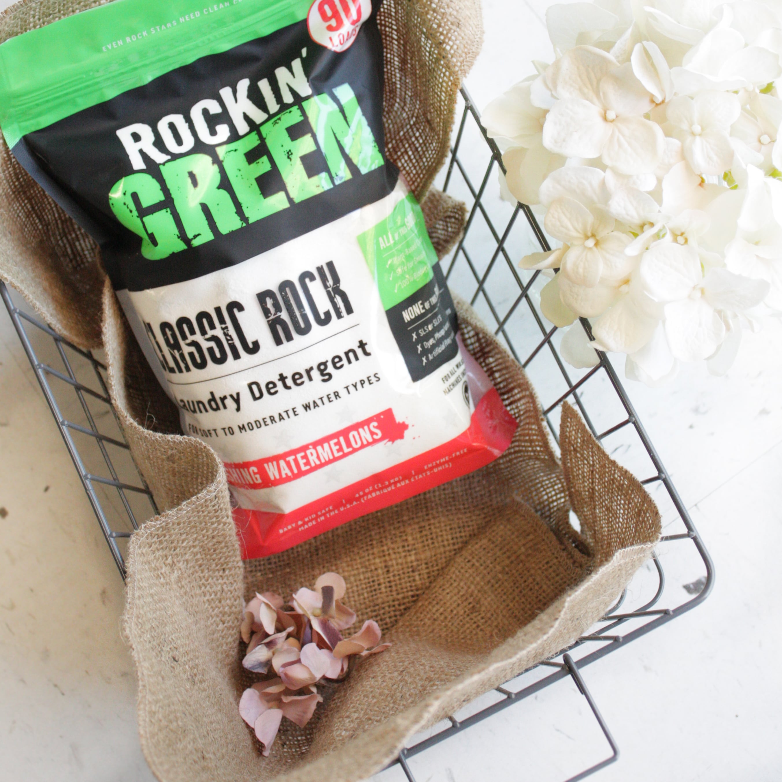 Rockin Green Classic Rock Detergent in a wire basket with burlap liner and white flowers nearby.